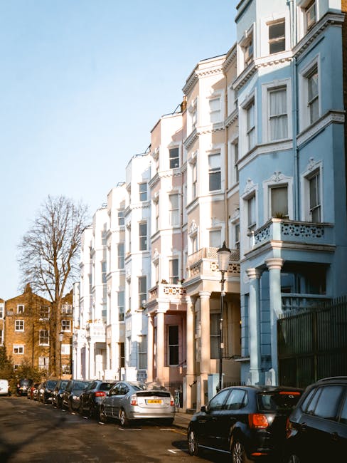 A row of Victorian-style residential buildings with ornate cornices and bay windows, painted in pastel shades of pink, white, and blue, situated on a street in Notting Hill. The buildings feature balc